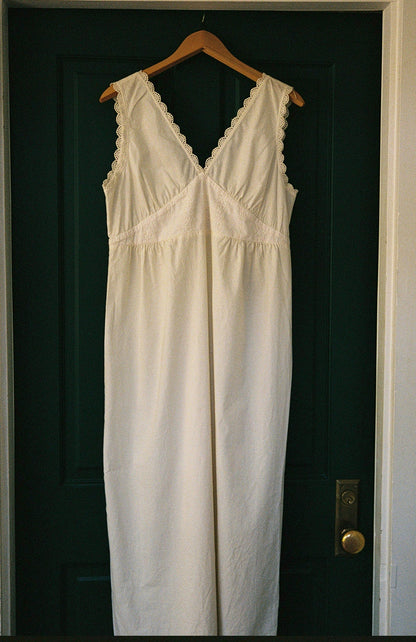 Butter - The Makinna Poplin Dress by LA RELAXED, a long sleeveless white organic poplin dress with delicate cotton lace on the straps and neckline, hangs on a wooden hanger against a dark green door.
