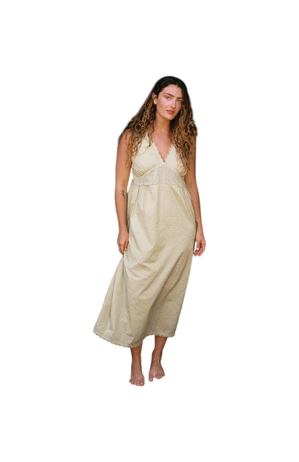 Butter - 
A woman with long wavy hair stands barefoot, facing forward in the LA RELAXED Makinna Poplin Dress—an ankle-length, sleeveless, light beige organic cotton dress with a V-neckline—embodying sustainable style against a plain white background.