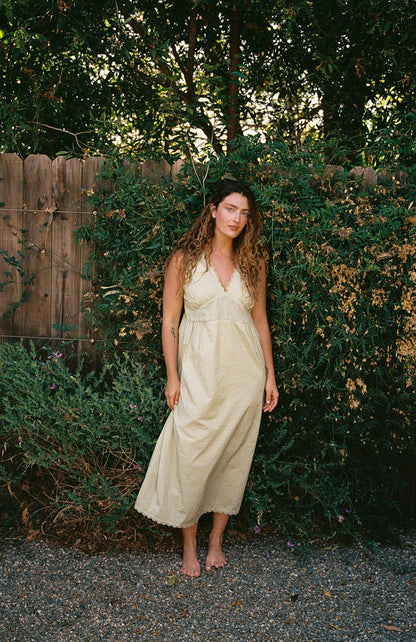 Butter - A woman with wavy hair stands barefoot on gravel in front of a wooden fence and green foliage, wearing the LA RELAXED Makinna Poplin Dress.