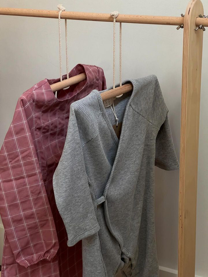 Two children's garments, a pink plaid dress and a cabane childrenswear Après Wrap Organic Waffle top in gray, hang on wooden hangers and rope from a minimalist wooden rack.