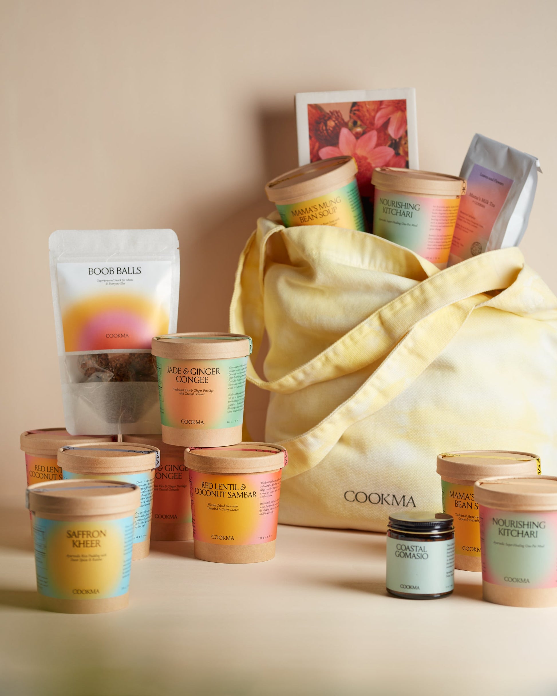 The Cookma Mama Bundle includes a yellow tote filled with vibrant food containers, teas, and snacks—ideal for postpartum care. Matching Cookma containers, nourishing meals, snacks, and two candles complete the set in front of the tote.