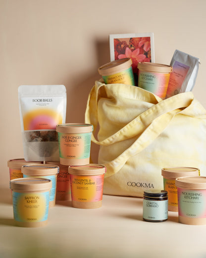 The Cookma Mama Bundle includes a yellow tote filled with vibrant food containers, teas, and snacks—ideal for postpartum care. Matching Cookma containers, nourishing meals, snacks, and two candles complete the set in front of the tote.