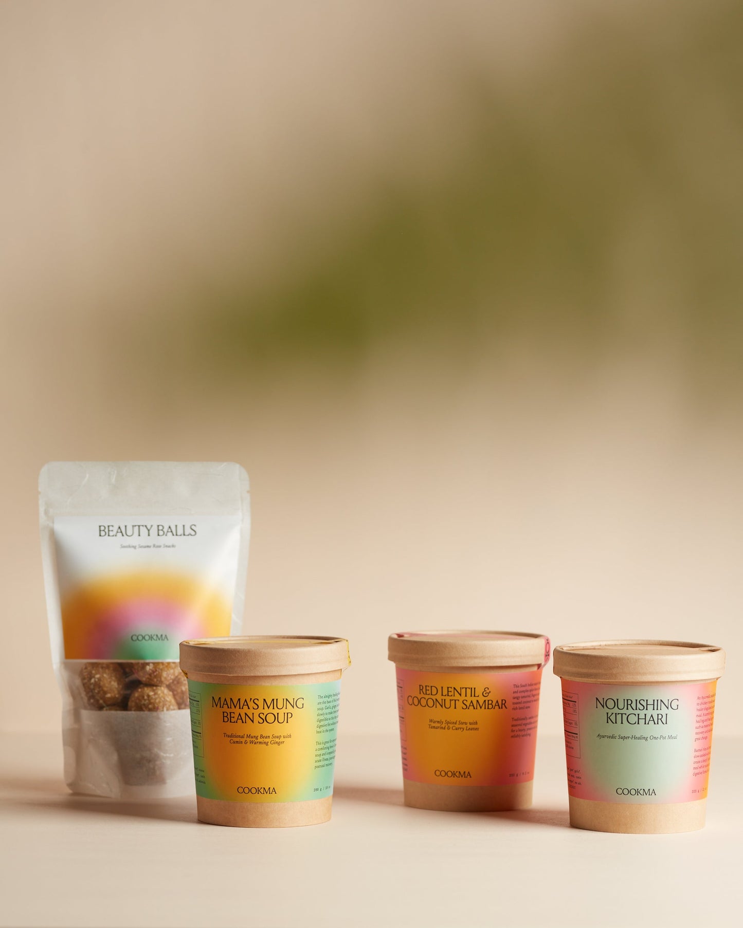 Four Cookma Wellness Bundle items—a Beauty Balls pouch and three tubs: Mama's Mung Bean Soup, Red Lentil & Coconut Sambar, and Nourishing Kitchari—offer comforting meals and snacks set against a neutral beige background.