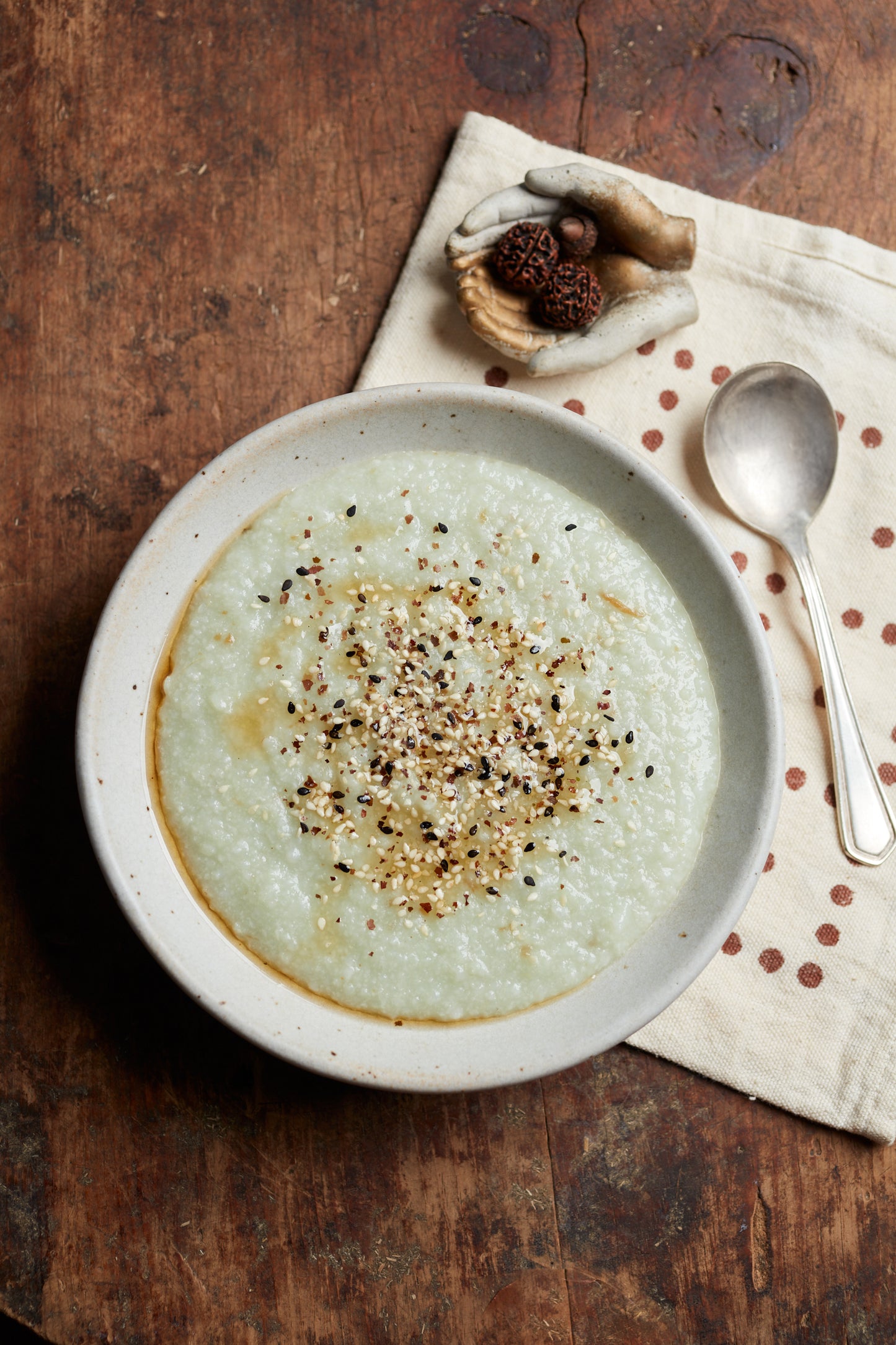 A bowl of Cookma’s Healing Bundle rice porridge, celebrated for its immune-supporting qualities, is garnished with seasoning and set on a beige napkin with brown dots, accompanied by a silver spoon and decor atop a rustic wooden table.