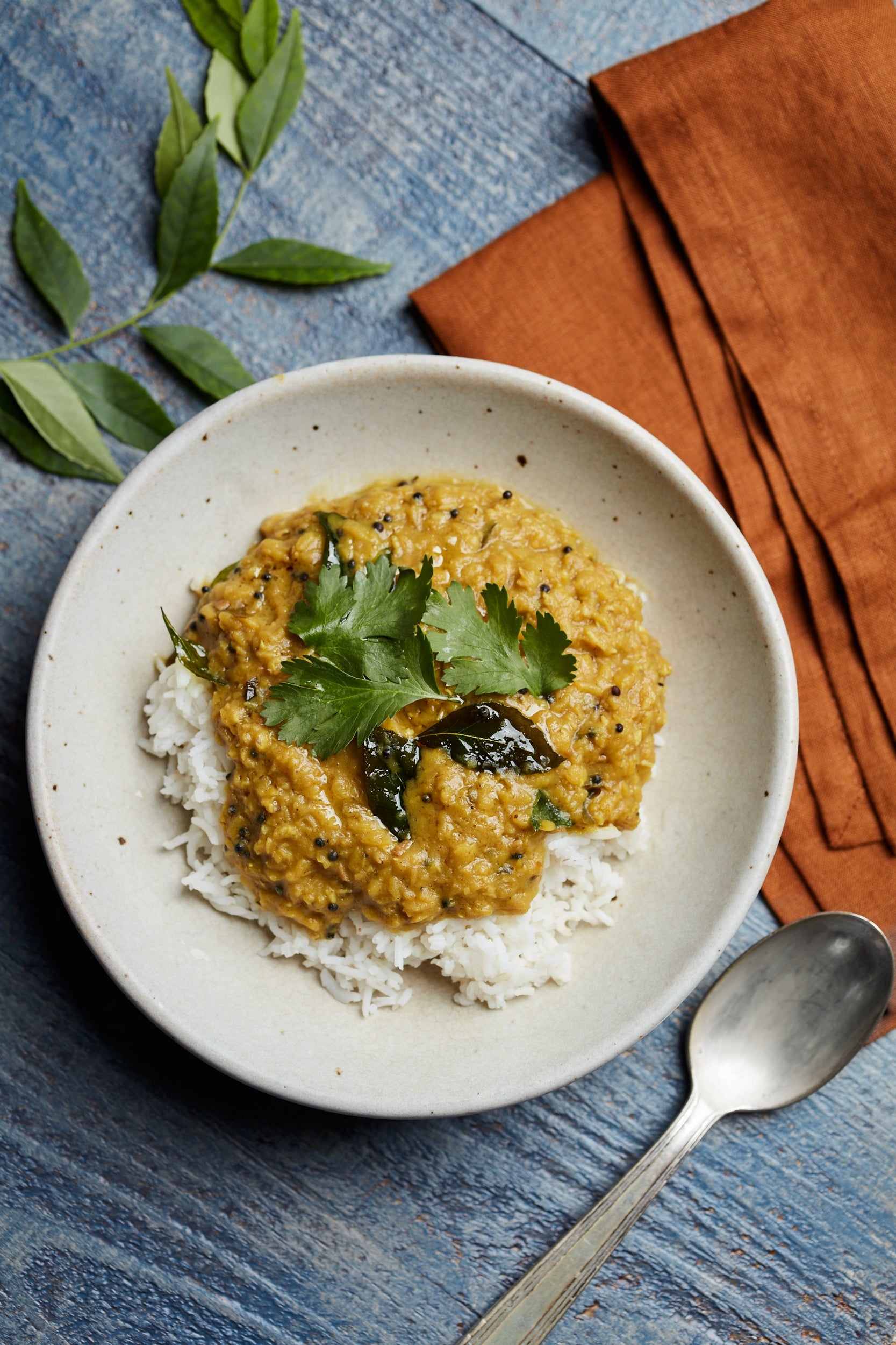A bowl of white rice with creamy yellow dal from Cookma’s Healing Bundle—an Ayurveda-inspired immune-boosting meal—is garnished with cilantro and curry leaves beside an orange napkin and silver spoon on a blue wooden surface.
