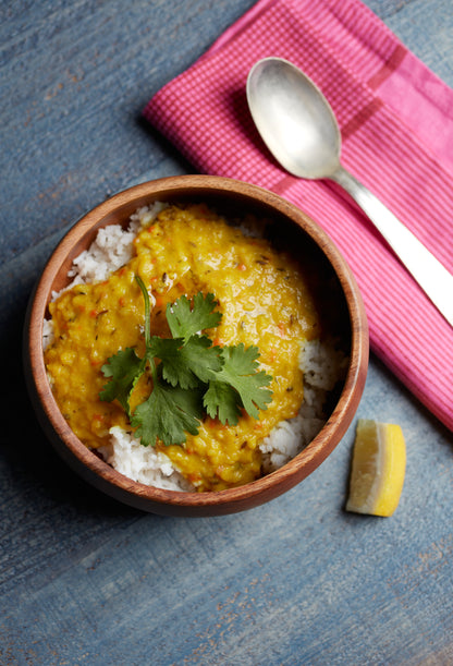 The Cookma Healing Bundle features a wooden bowl of white rice topped with yellow dal and cilantro, set on a blue surface with a lemon slice, spoon, and pink cloth napkin—Ayurveda-inspired nourishment at its finest.