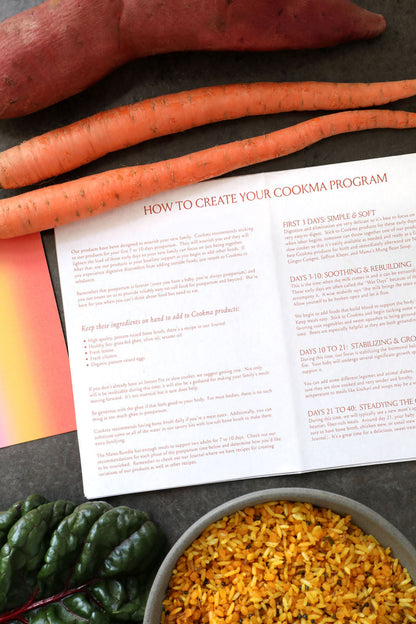 The Cookma Mama Bundle booklet, titled "How to Create Your Cookma Program," rests on a table with carrots, sweet potato, greens, and yellow rice. This guide offers nourishing meals and meal phase instructions for postpartum support.
