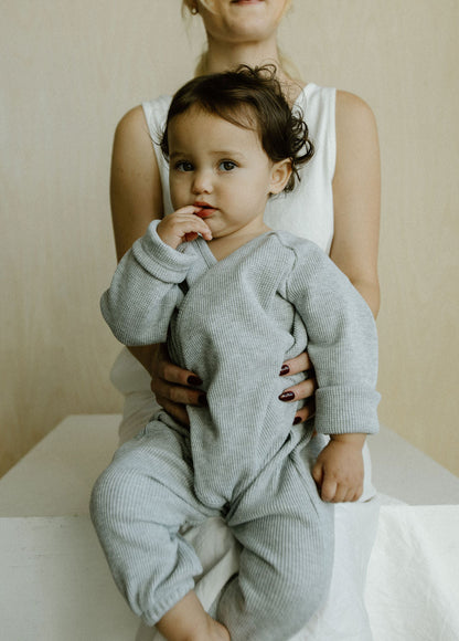 A toddler in an Après Wrap Organic Waffle onesie by cabane childrenswear sits on a woman's lap, partially visible in a white outfit. 