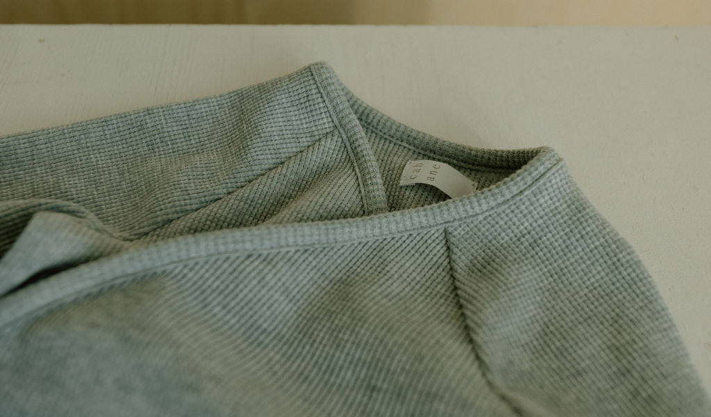 Close-up of the Après Wrap Organic Waffle by cabane childrenswear, a light green textured sweater, showcasing its white label. Ethically handmade from organic cotton waffle knit, it rests on a light surface, slightly folded to highlight its cozy design.