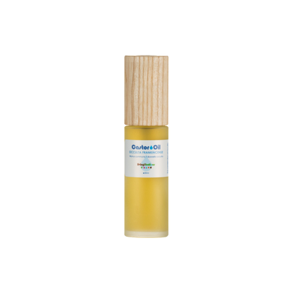 A small cylindrical gold bottle with a light wooden cap, containing cold-pressed Castor Oil rich in ricinoleic acids, is labeled "Castor Oil + Occulta Frankincense." It features a small white label with colorful text, set against a plain black background. The brand is Living Libations.