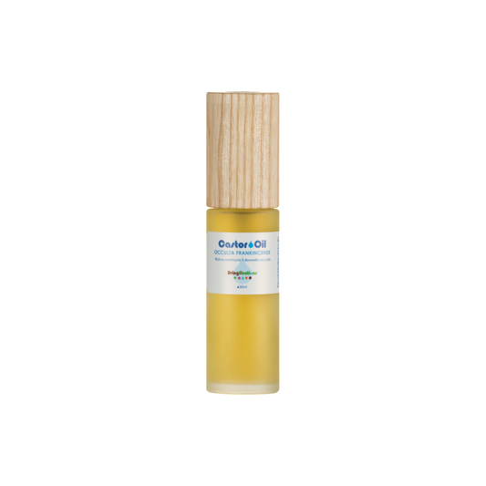 A small cylindrical gold bottle with a light wooden cap, containing cold-pressed Castor Oil rich in ricinoleic acids, is labeled "Castor Oil + Occulta Frankincense." It features a small white label with colorful text, set against a plain black background. The brand is Living Libations.