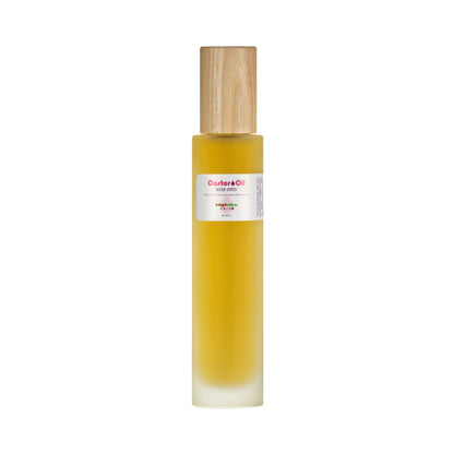 The tall, cylindrical bottle of Living Libations' Castor Oil + Rose Otto has a wooden cap and features a clear label with text and color accents against the golden backdrop of ricinoleic acid-rich oil. It stands elegantly against a plain white background.