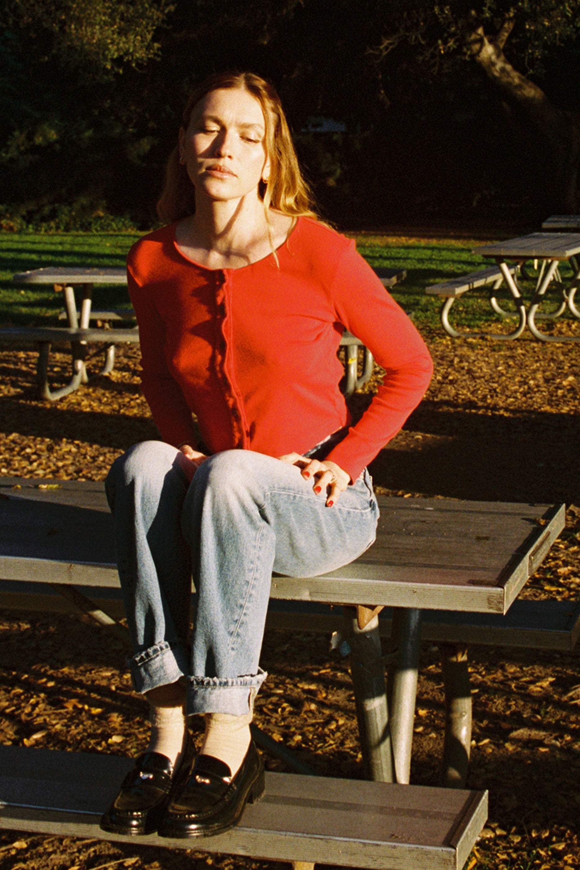 Cherry 
A woman wearing an Organic Rib Cardigan from LA Relaxed sits cross-legged on a picnic table in jeans, her eyes closed in relaxation. The scene is sunlit with greenery and more tables around, creating a warm 90's closet vibe.