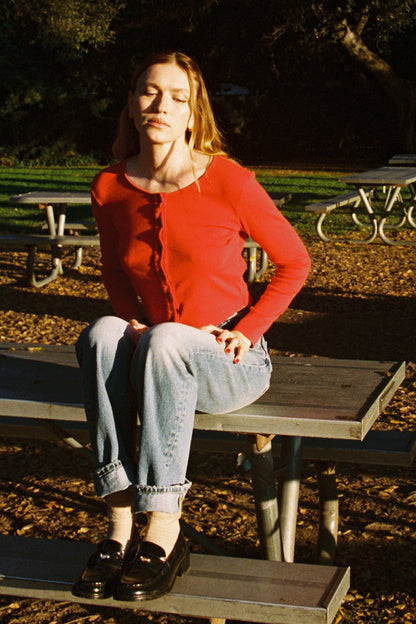 Cherry 
A woman wearing an Organic Rib Cardigan from LA Relaxed sits cross-legged on a picnic table in jeans, her eyes closed in relaxation. The scene is sunlit with greenery and more tables around, creating a warm 90's closet vibe.