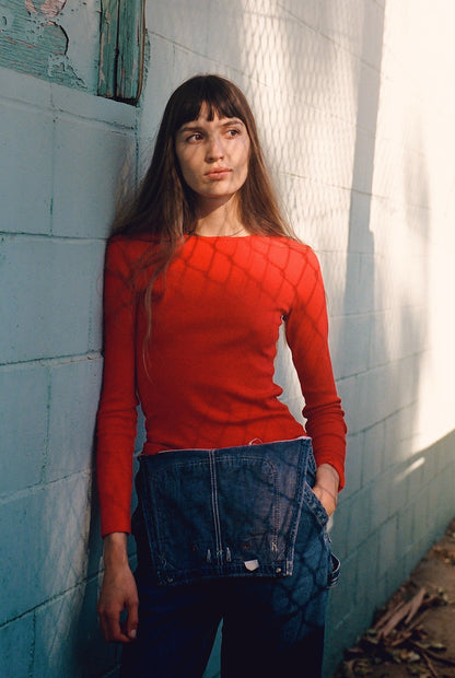 A woman in a Bebe Organic Long Sleeve by LA RELAXED leans against a light blue wall holding a denim jacket. Shadows of a chain-link fence fall across her and the wall, with leaves scattered on the ground.
