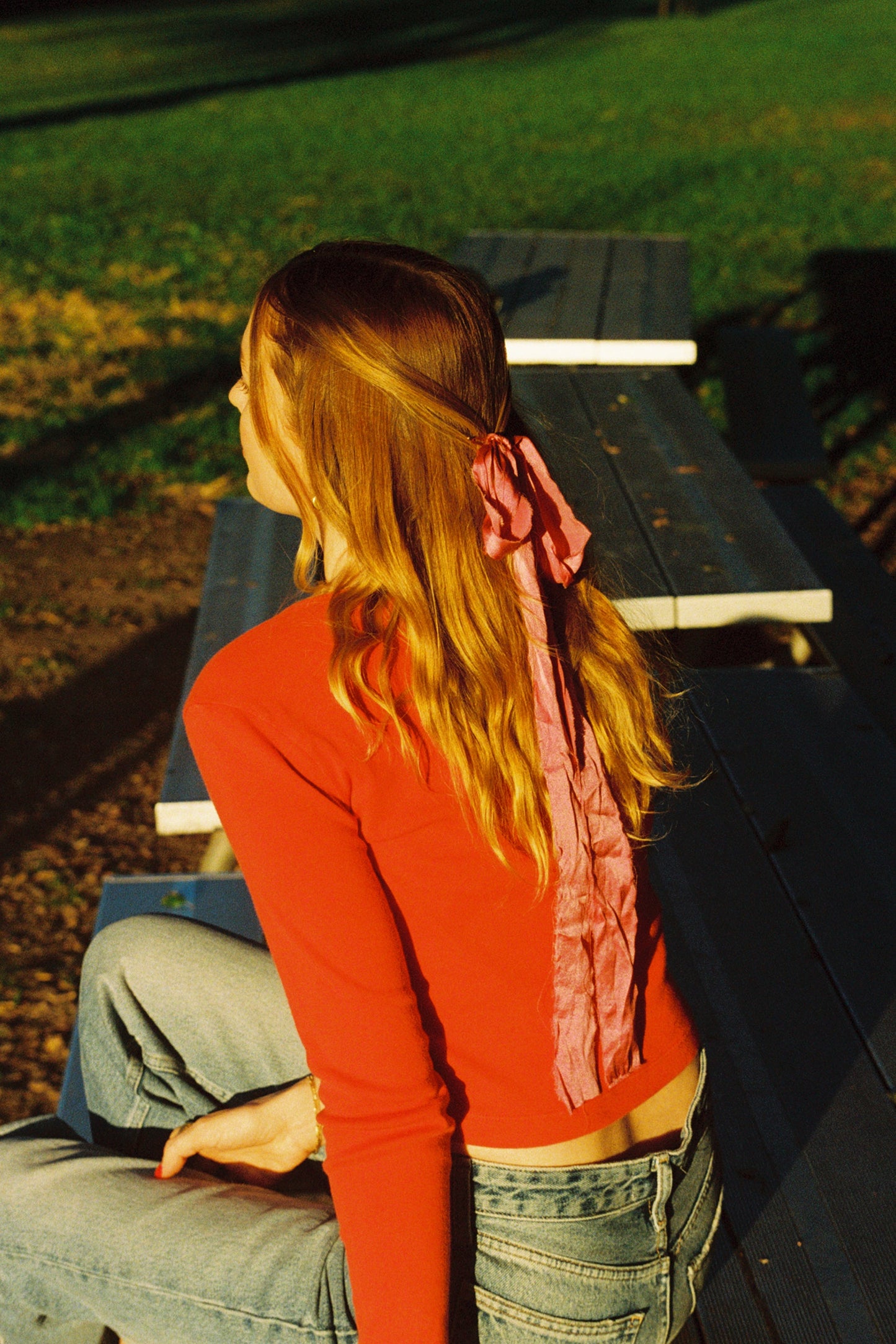 Cherry 
A person with long, wavy hair sits sideways on a picnic table in the park, wearing a red top and jeans. A pink ribbon adorns their hair as they savor the sunlight, casually draping an LA Relaxed Organic Rib Cardigan over their shoulder.