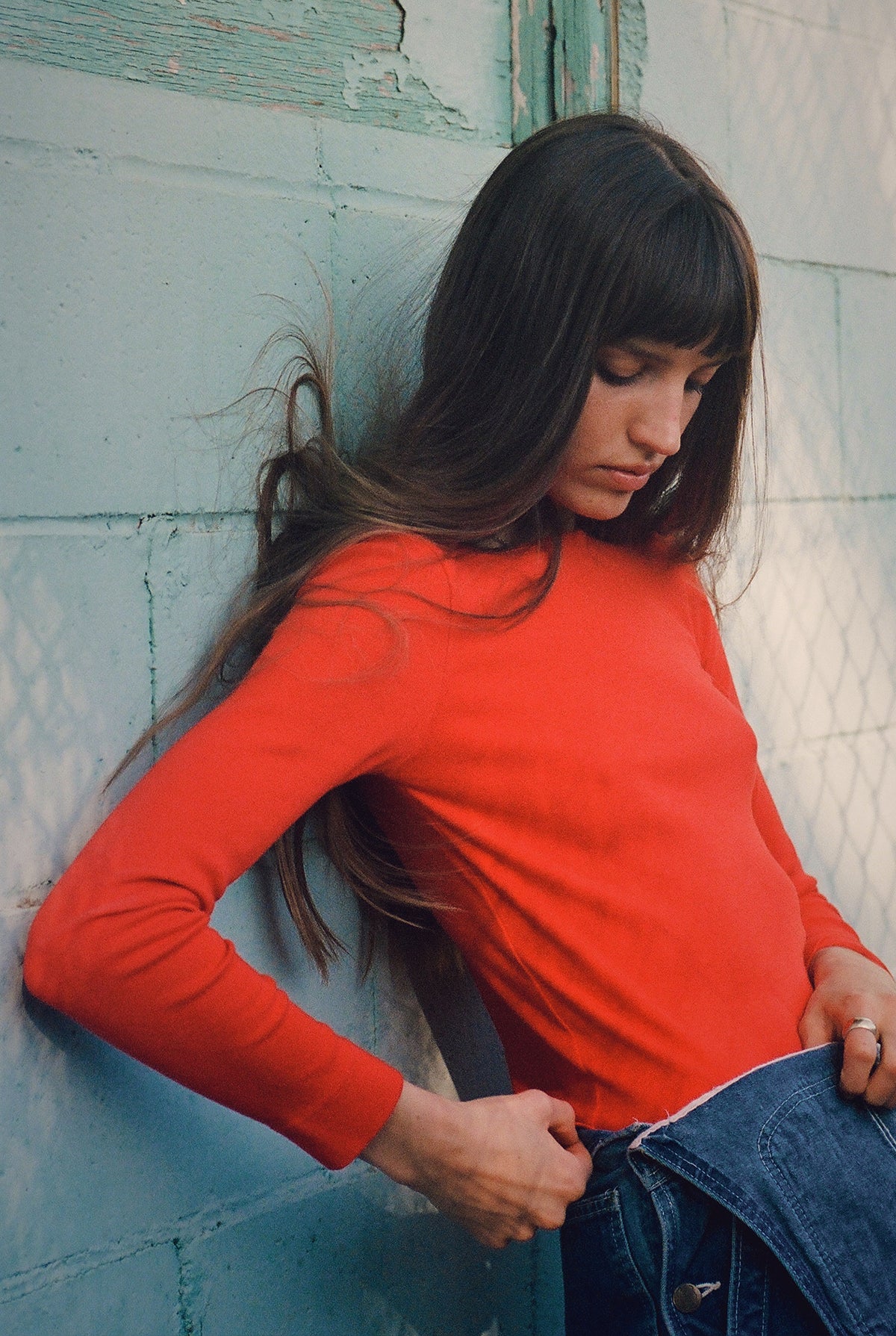 A person with long brown hair and bangs leans against a teal brick wall wearing the LA RELAXED Bebe Organic Long Sleeve in red, paired with blue jeans. They look down, one hand resting in their pocket.