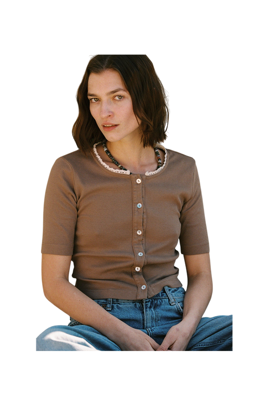 A person with shoulder-length brown hair is seated, looking forward. They're wearing an LA Relaxed Short Sleeve Organic Cardi, paired with blue jeans against a plain white backdrop.