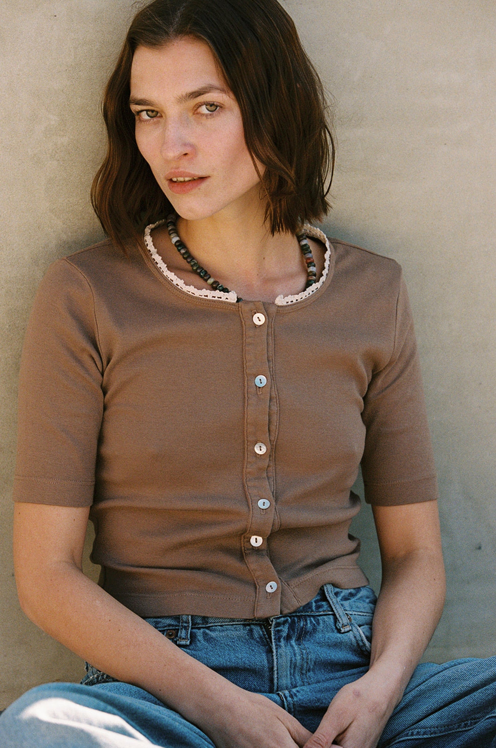 A person with shoulder-length brown hair sits against a plain background, wearing LA Relaxed's Short Sleeve Organic Cardi with shell buttons and jeans. They have a neutral expression and wear a beaded necklace.