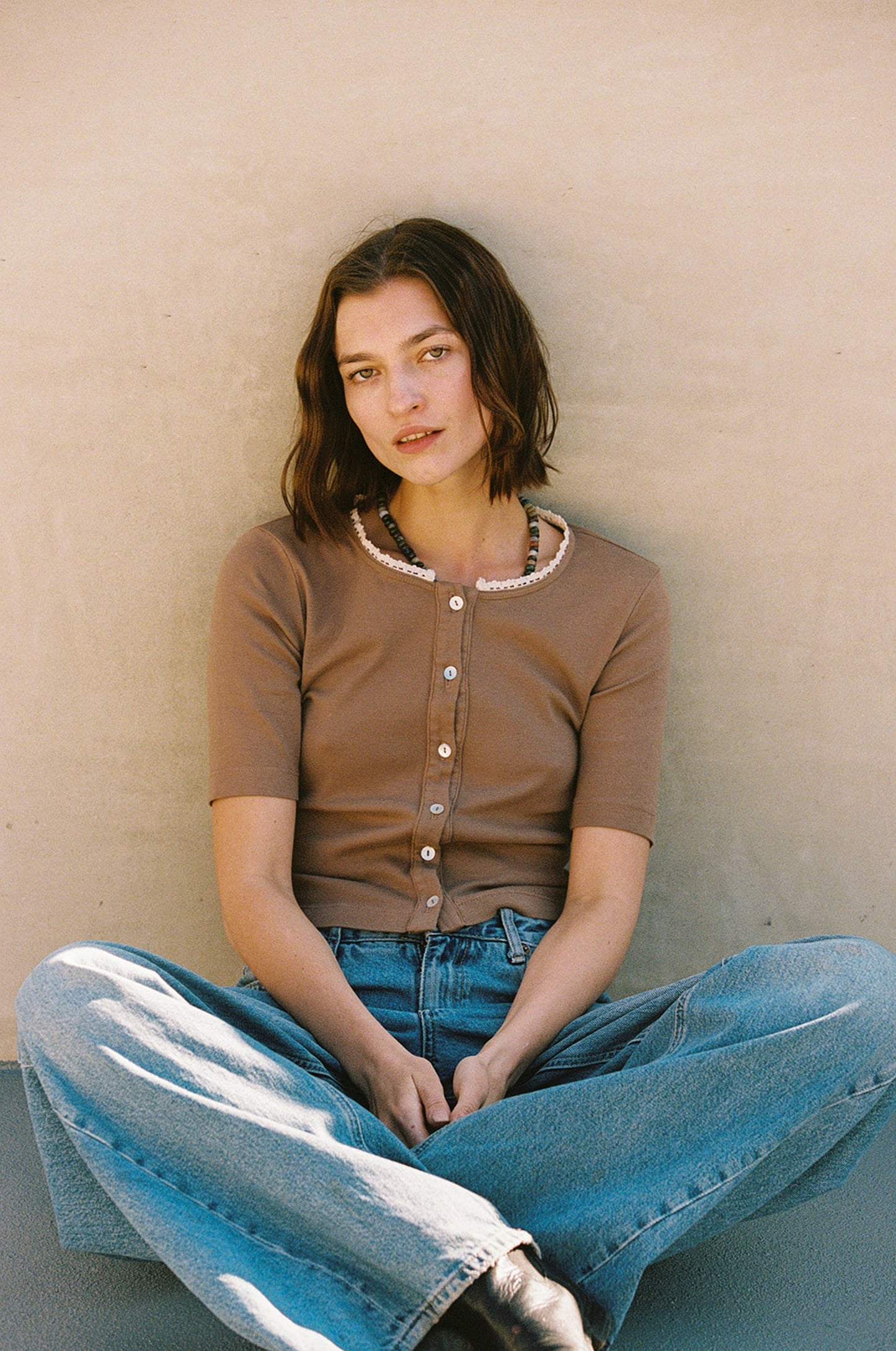 A woman with shoulder-length brown hair leans against a beige wall, wearing LA Relaxed's Short Sleeve Organic Cardi in brown with shell buttons and a pearl necklace. She pairs it with lace-trimmed blue jeans and rests her hands on her lap with a relaxed expression.
