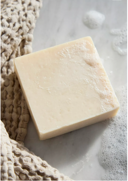 A square Collagen Tallow Facial Bar by My Neighbor’s Tallow, in light beige with suds, sits on a white surface beside a textured tan towel.