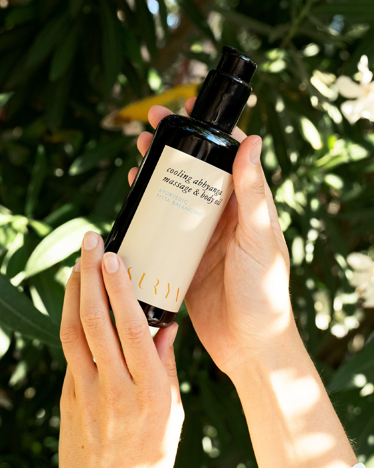 In an image, two hands gently hold a dark bottle of Surya's Cooling Body Oil against a backdrop of lush green leaves. The bottle, capped in black and adorned with elegant typography, is designed to balance Pitta dosha with its soothing properties.