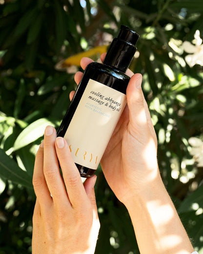 In an image, two hands gently hold a dark bottle of Surya's Cooling Body Oil against a backdrop of lush green leaves. The bottle, capped in black and adorned with elegant typography, is designed to balance Pitta dosha with its soothing properties.