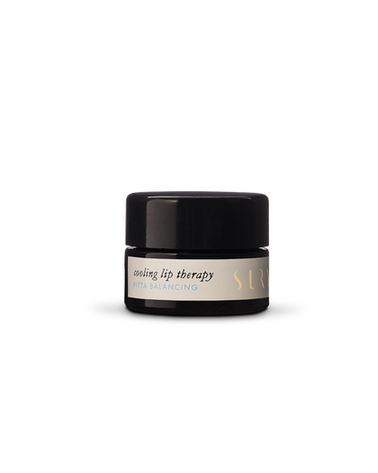 A small black cylindrical container of Cooling Lip Therapy, labeled "Pitta Balancing" in gold and white text on a light-colored background, with the brand name "Surya" partially visible. This lip balm features natural blends creating an organic ingredient base for soothing relief.