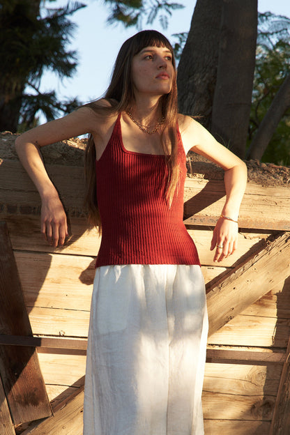 Cranberry A woman stands outdoors in sunlight, leaning against a rustic wooden fence. She wears the LA Relaxed Pointelle Square Cami in red with a long skirt; trees and greenery fill the background.