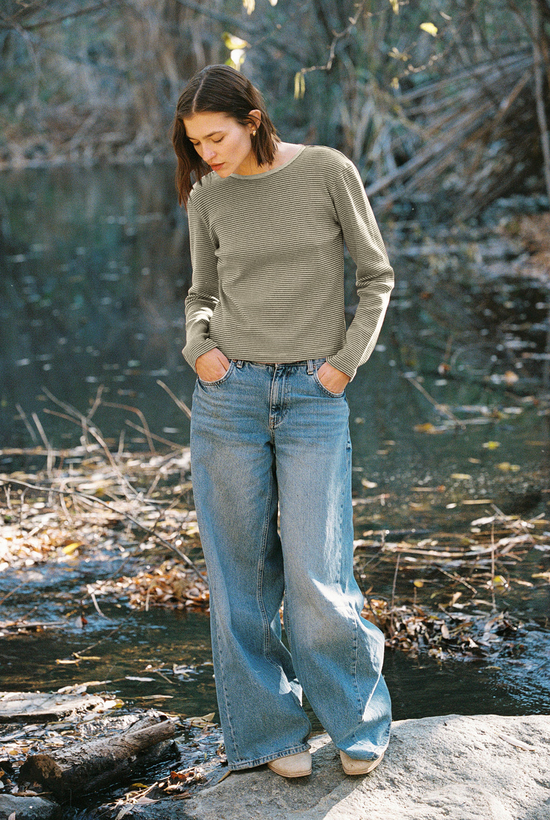 A person with shoulder-length brown hair, wearing an Organic Stripe Long Sleeve by LA RELAXED and wide-leg blue jeans, stands on a rock near a serene river surrounded by trees and fallen leaves.