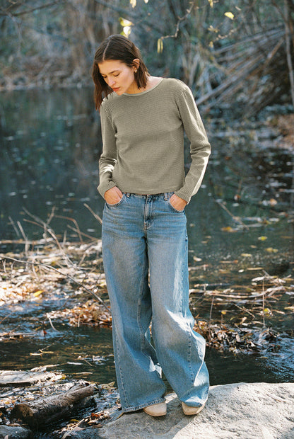 A person with shoulder-length brown hair, wearing an Organic Stripe Long Sleeve by LA RELAXED and wide-leg blue jeans, stands on a rock near a serene river surrounded by trees and fallen leaves.