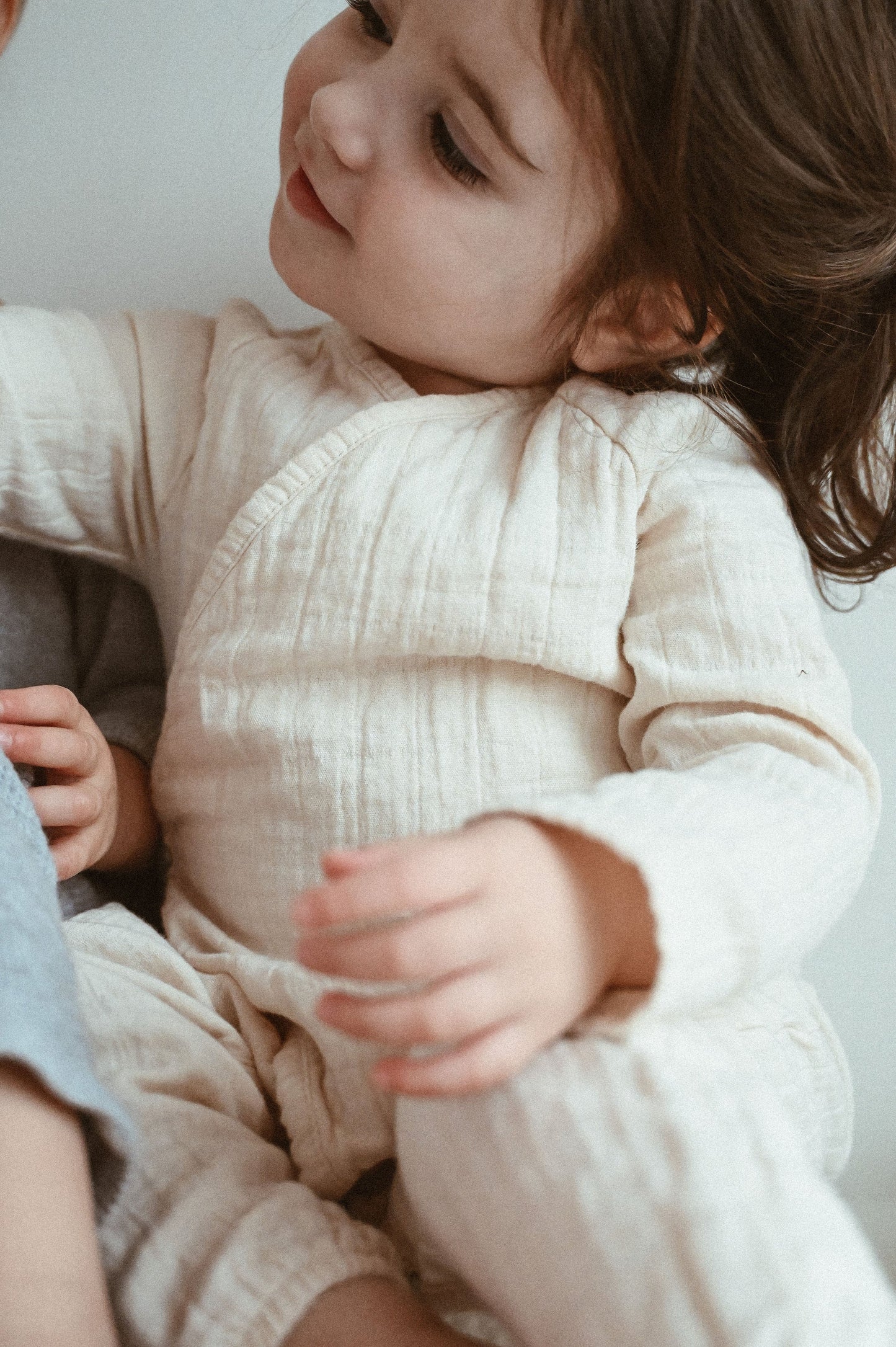 A young child with brown hair gazes to the side while wearing a soft, cream-colored outfit made from breathable fabric. The child is sitting with a relaxed, gentle expression against a neutral background, wrapped in the comfort of the Après Organic Wrap Undyed by cabane childrenswear.