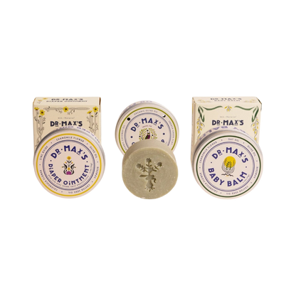 The Dr. Max's Baby Essentials Bundle features vintage-style packaging: two boxes and tins for Baby Balm and Diaper Ointment, plus a round Olive Oil Soap bar with a floral imprint nestled between them.
