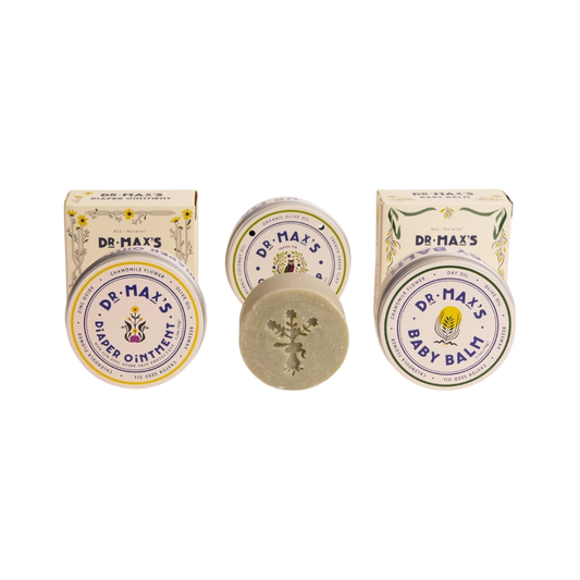 The Dr. Max's Baby Essentials Bundle features vintage-style packaging: two boxes and tins for Baby Balm and Diaper Ointment, plus a round Olive Oil Soap bar with a floral imprint nestled between them.