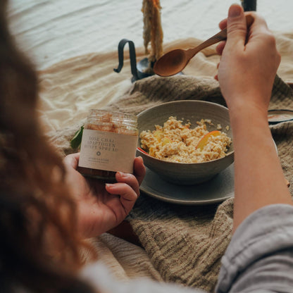 Person holding a jar of "Rose Chai Adaptogen Honey" by Moon Canyon Healing and a wooden spoon, in front of a bowl of oatmeal on a table adorned with a textured cloth. The scene exudes coziness with its warm, natural ambiance, emphasizing the benefits of adaptogenic honey for promoting healthy circulation.