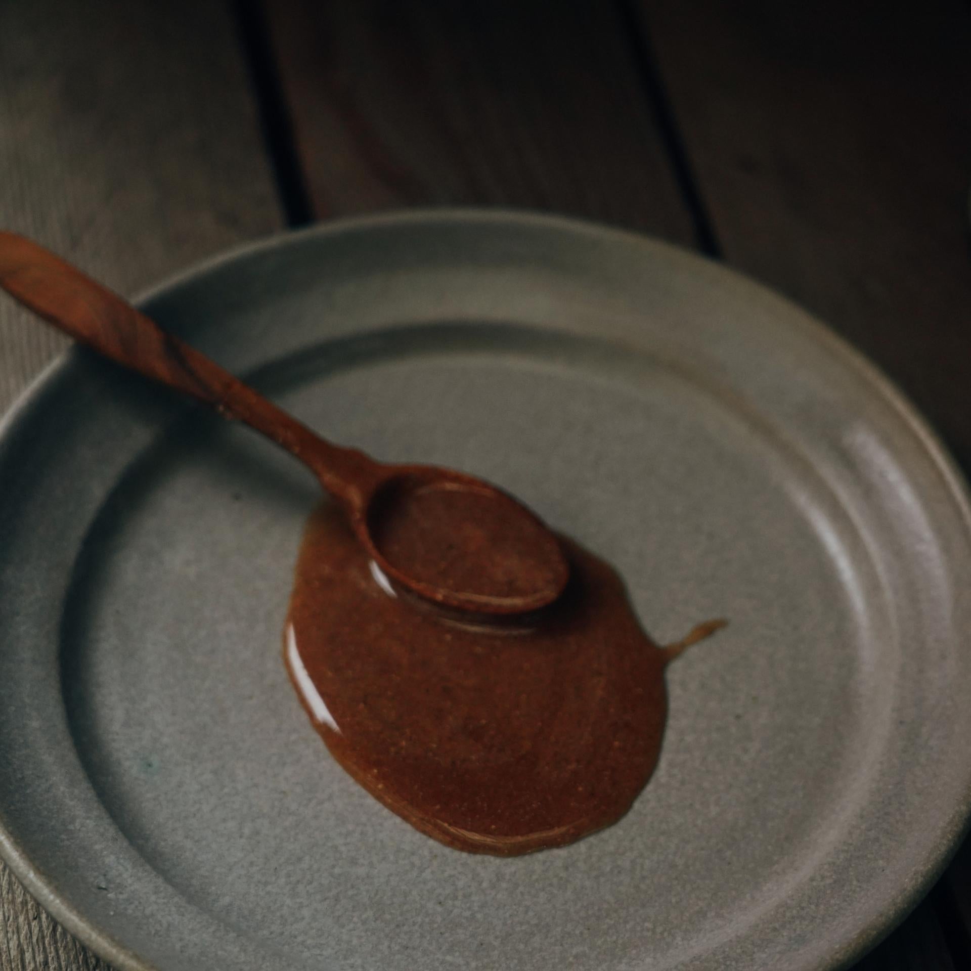 A grey ceramic plate holds a wooden spoon coated in Moon Canyon Healing's Rose Chai Adaptogen Honey, its rich, chai-spiced liquid dripping onto the plate. The dimly lit scene, set against a rustic wooden background, exudes warmth and charm.