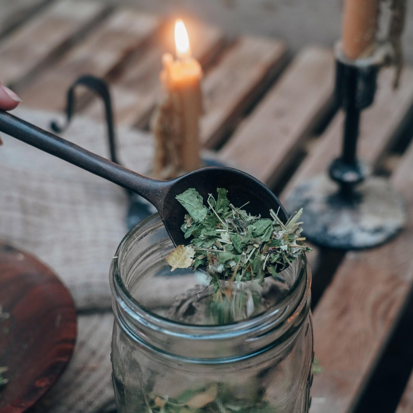 A hand holds a spoon over a glass jar filled with Fill The Well Herbal Infusion from Moon Canyon Healing, creating a calming herbal brew, while lighting in the background comes from two lit candles on a wooden table. The scene embodies warm, rustic self care.