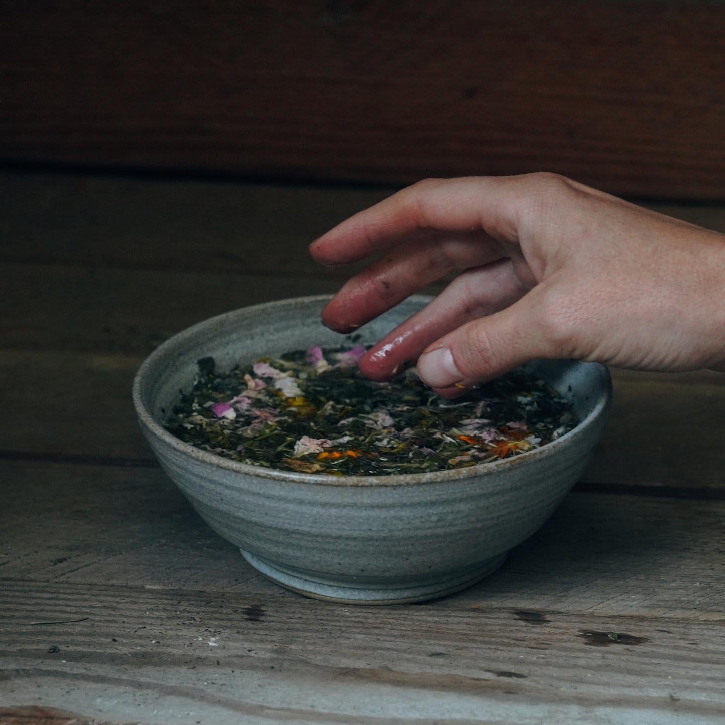 A hand reaches into a ceramic bowl filled with Moon Canyon Healing's Sacred Steam Herbal Yoni Blend, a mixture of chopped herbs and colorful petals. The bowl is set on a rustic wooden surface, with the background blurred to focus attention on the hand and the blend.