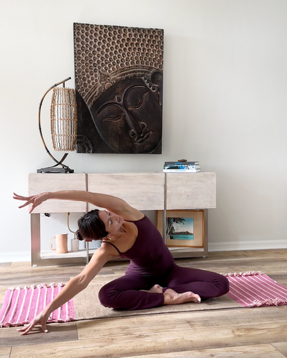 A woman in a purple yoga outfit stretches gracefully on a Bennd Yoga Dark Harda and Sappan Wood Ayurvedic Yoga Mat in her living room. Behind her, a wooden sideboard holds a lamp and books, while a large Buddha artwork adorns the wall. The floor is wooden with pink-striped rugs, casting a serene ambiance throughout the space.