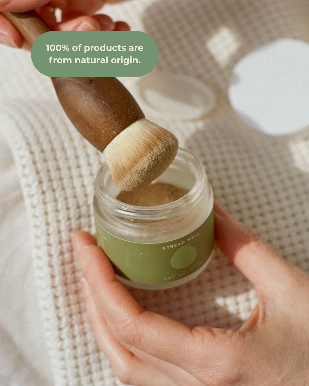 A hand dips an & Tread Well Dry Shampoo wooden brush into a small jar with a green label. Above, text reads, "100% of products are from natural ingredients." The background features textured cream-colored fabric.