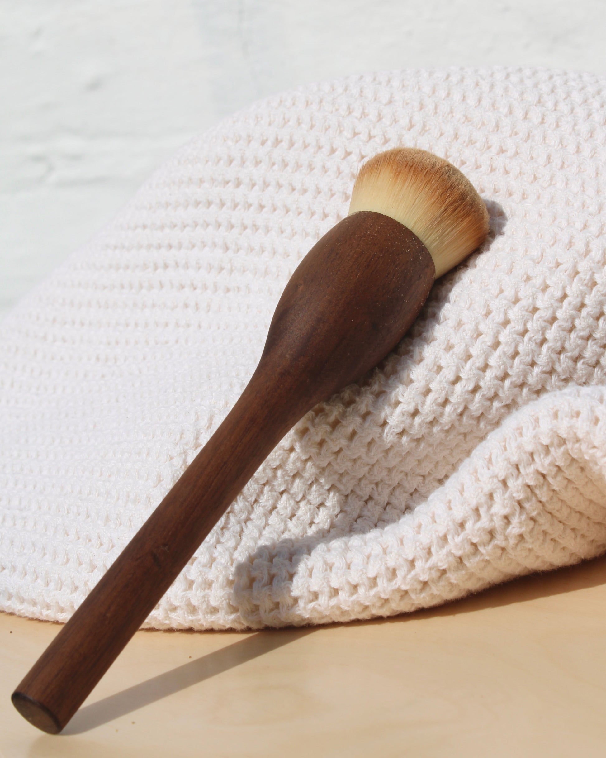The & Tread Well Dry Shampoo Brush, featuring a wooden handle and soft natural bristles, rests on a cream-colored waffle towel atop a light wooden surface with a white background.