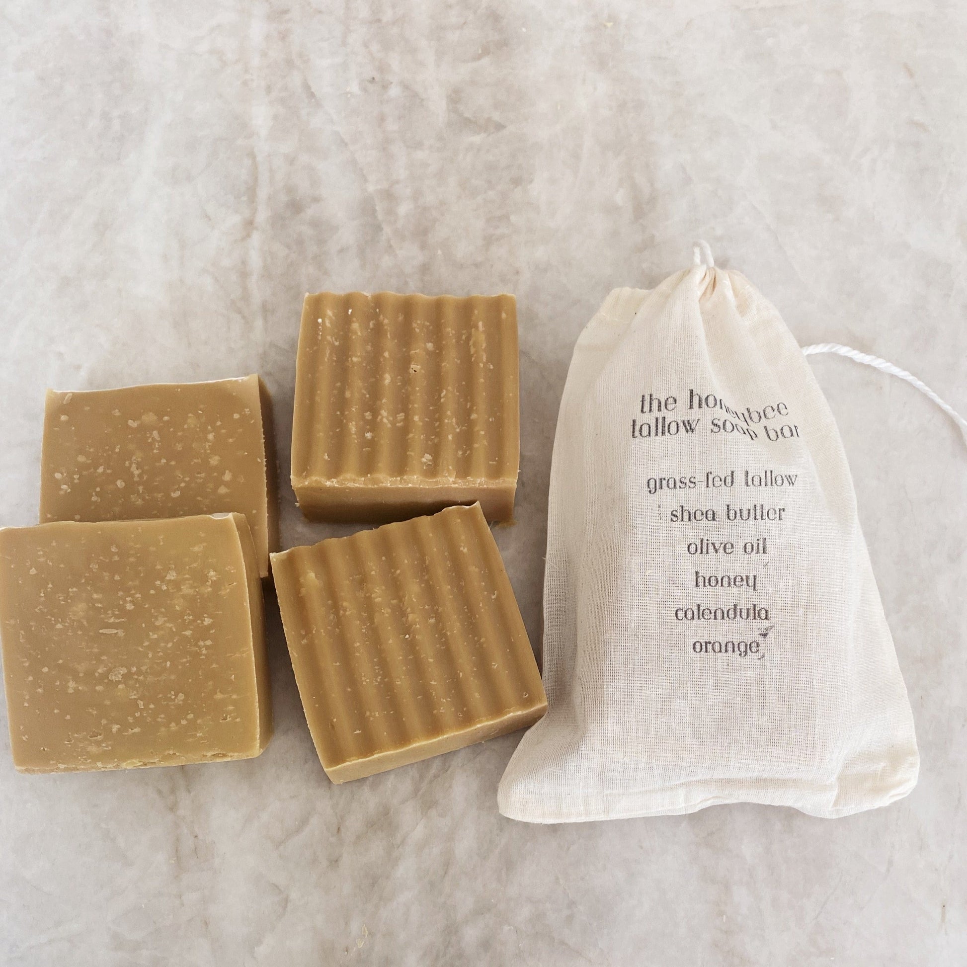 Two wavy and two smooth handmade 'the honeybee tallow soap bars' by Honeybee Hippie sit alongside a drawstring bag. The bag lists ingredients like grass-fed tallow, shea butter, honey, and organic herbs on a light textured surface.