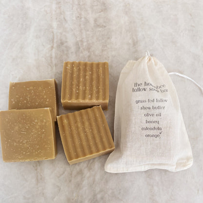 Two wavy and two smooth handmade 'the honeybee tallow soap bars' by Honeybee Hippie sit alongside a drawstring bag. The bag lists ingredients like grass-fed tallow, shea butter, honey, and organic herbs on a light textured surface.