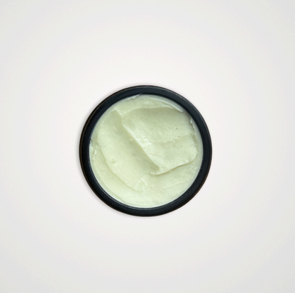 A round, black container of Wild Mamas Earth, Whipped & Sky 3.16oz tallow balm filled with smooth, pale green whipped balm, viewed from above against a plain white background.