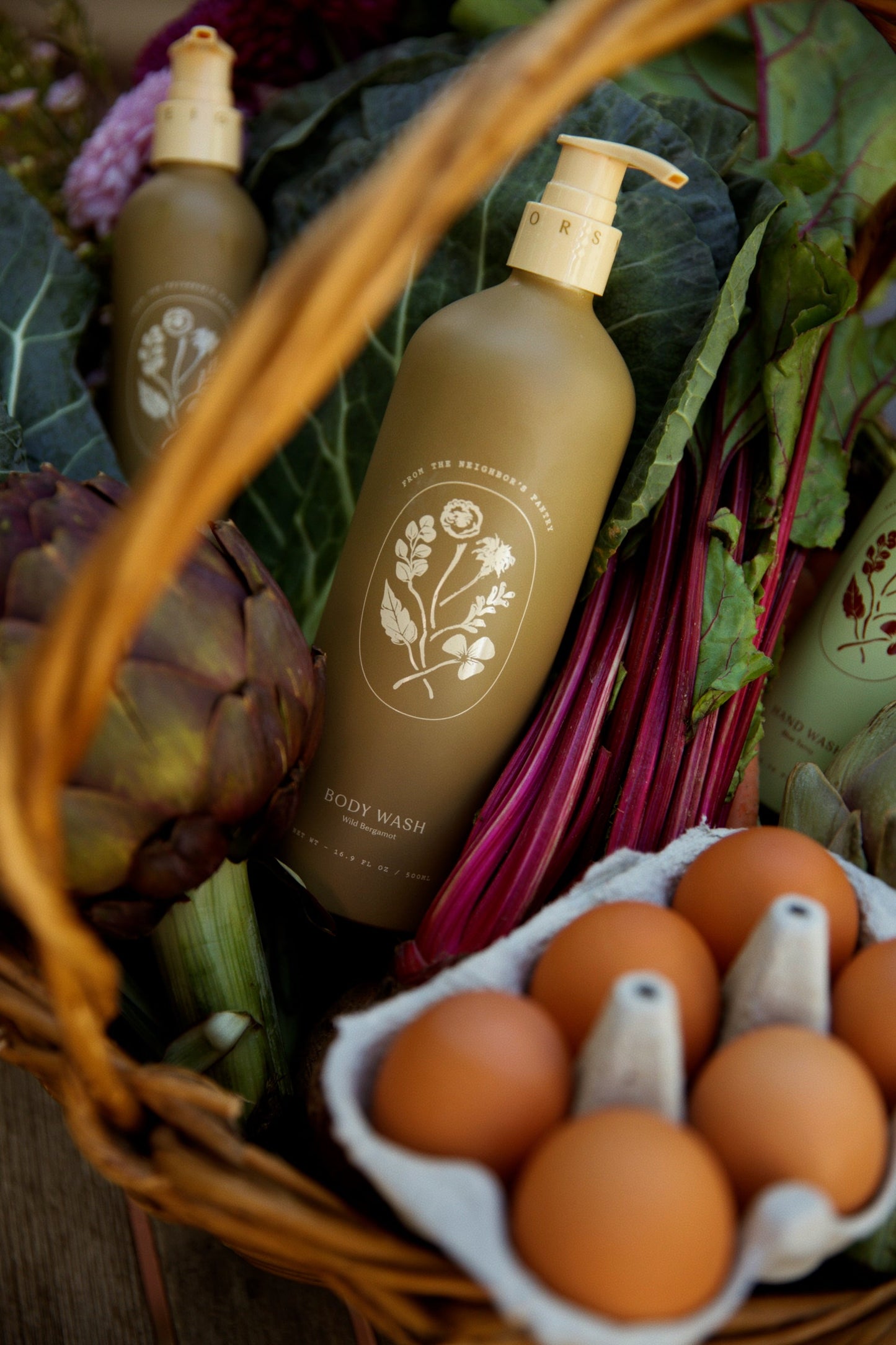 A basket with My Neighbor’s Tallow Wild Bergamot Body Wash (floral label), a carton of brown eggs, an artichoke, leafy greens, and other non-GMO vegetables.
