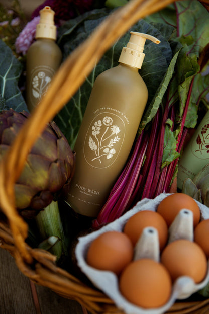 A basket with My Neighbor’s Tallow Wild Bergamot Body Wash (floral label), a carton of brown eggs, an artichoke, leafy greens, and other non-GMO vegetables.
