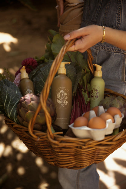 A person holds a wicker basket with leafy greens, an artichoke, brown eggs, and two My Neighbor’s Tallow Wild Bergamot Body Wash bottles featuring botanical labels and non-GMO ingredients. Their arm, in denim and a bracelet, is visible.