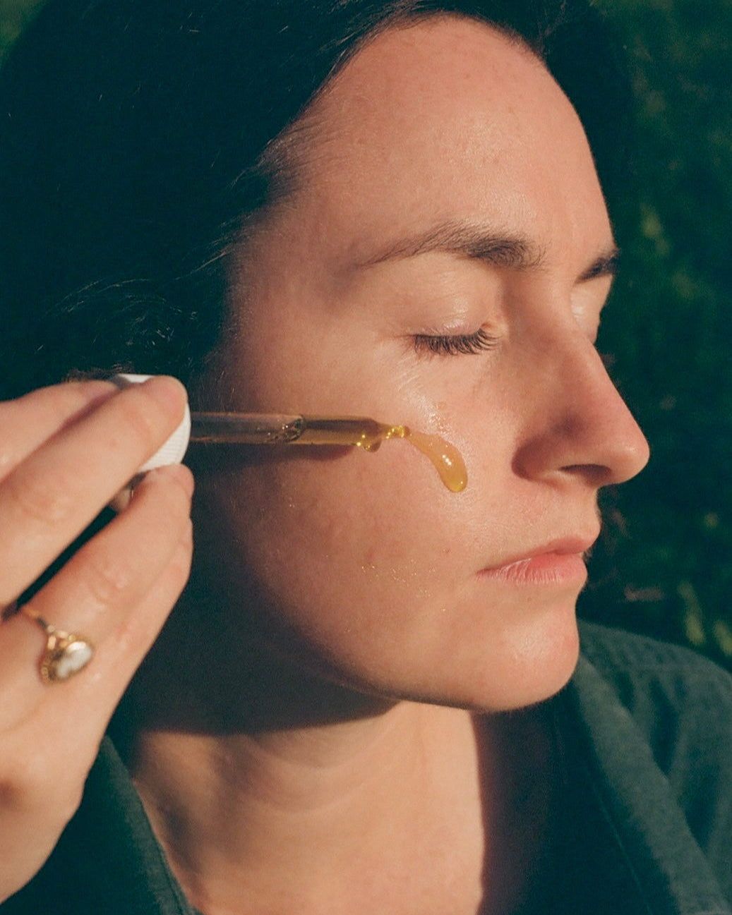With eyes closed, a person applies & Tread Well Face Serum to their cheek using a dropper. Sunlight highlights their face, and they wear a gold ring featuring a heart-shaped design.