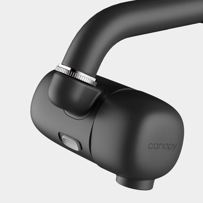 Close-up of the Canopy Faucet Filter, a sleek black device with a dial and button, attached to a faucet. "Canopy" is printed on the side. The background is plain light gray. Fulfilled by Canopy.