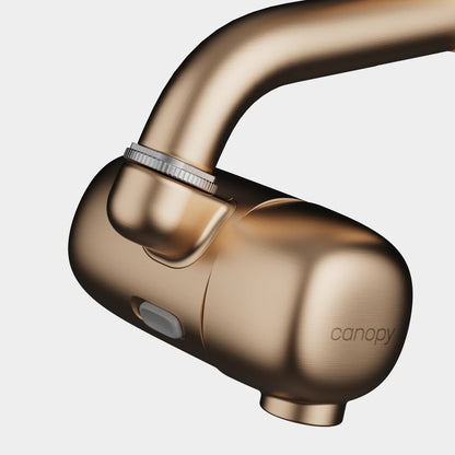 A close-up of the Canopy Faucet Filter, a modern metallic bronze attachment with "canopy" engraved on the side, featuring a smooth curved design and a small front button. Fulfilled by Canopy.